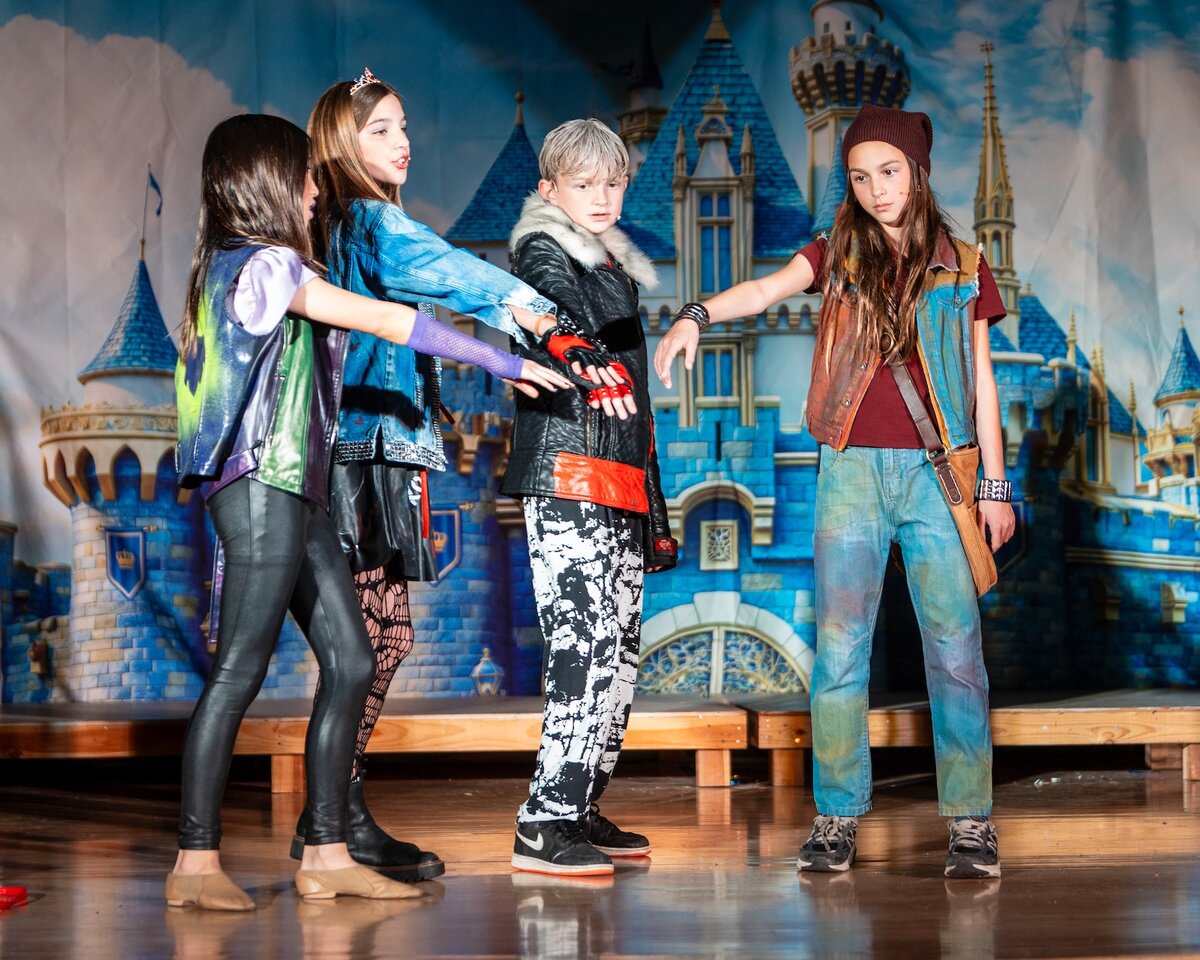 Student cast members in Descendants costumes performing a dance number on stage in front of a castle backdrop